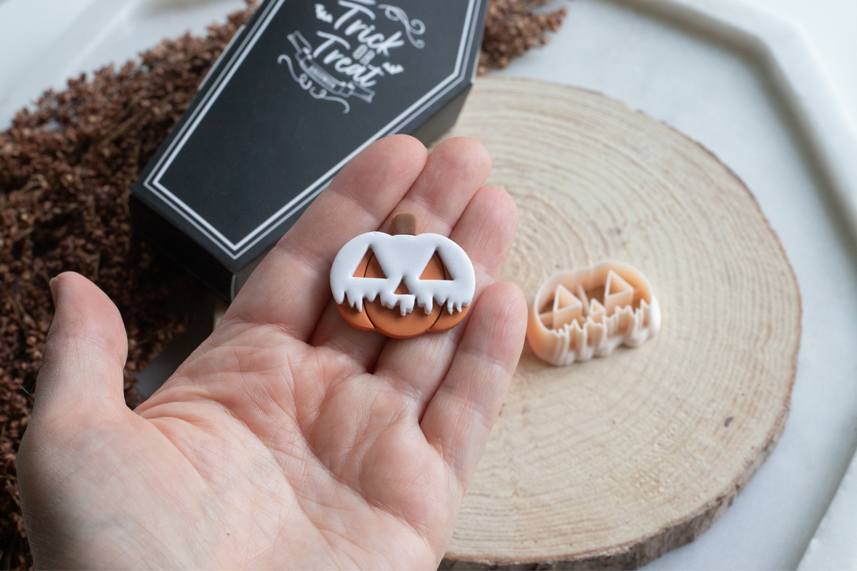 A hand holds a small jack-o'-lantern-shaped Halloween treat or charm, with a matching piece resting on a wood slice in the background. A black coffin-shaped box labeled 'Trick or Treat' is also visible on a white tray.