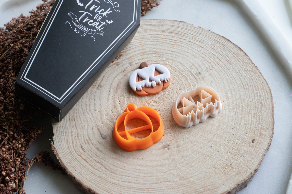 Three Halloween-themed cookie cutters in orange and beige are displayed on a round wood slice next to a black coffin-shaped box with white script that reads 'Trick or Treat'.