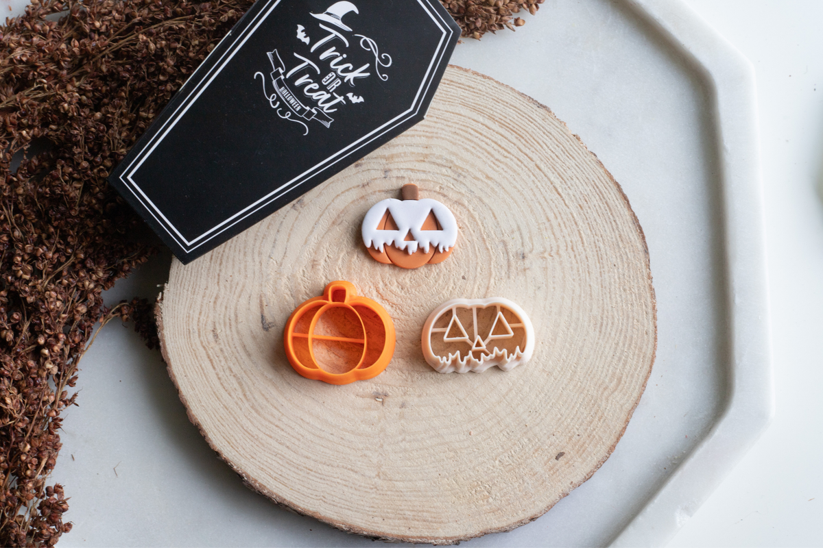 Two Halloween-themed Clay cutters displayed on a round wood slice with a black coffin-shaped tag that reads 'Trick or Treat'. The cutters include a jack-o'-lantern face, a pumpkin body, and a triangular pumpkin head design, shown on a light gray surface with decorative dried foliage.