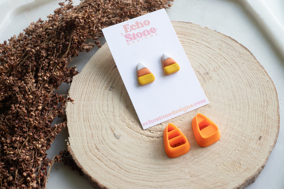 A pair of small candy corn stud earrings on a white Echo Stone Designs display card, with matching orange and yellow candy corn earrings placed below on a round wood slice. Dried brown flowers sit to the left on a white surface.