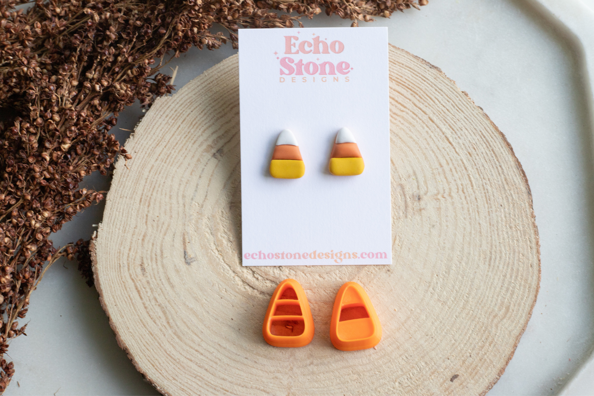 Candy corn and orange stud earrings displayed on a white backing card labeled Echo Stone Designs, arranged on a round wood slice with dried brown botanicals in the background.