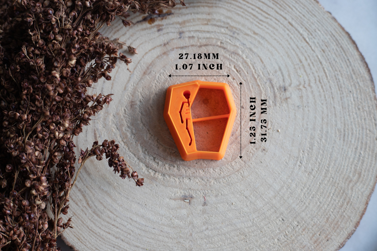 An orange hexagonal Clay cutter with a skeleton design, displayed on a light wood slice with dried flowers and dimension lines showing its measurements.