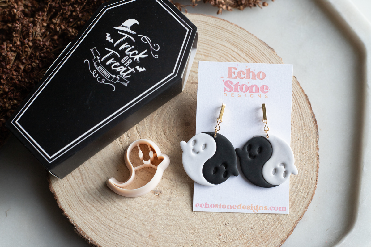 Black and white yin-yang style ghost drop earrings displayed on a white card labeled Echo Stone Designs, with a small peach-colored ghost stud and a black coffin-shaped box on a wood slice background.