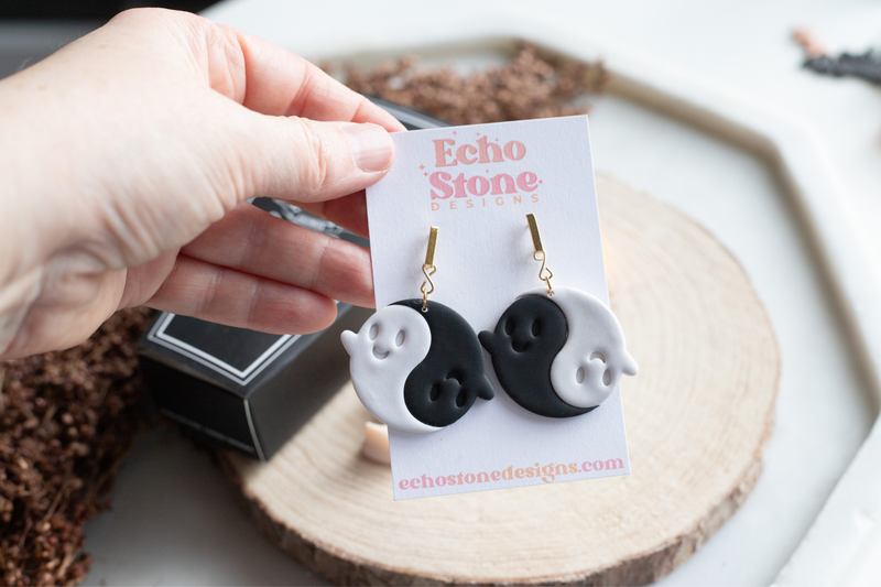 A hand holds a pair of black and white ghost yin-yang earrings on a white display card branded Echo Stone Designs, with a wooden slice and dark box in the background.