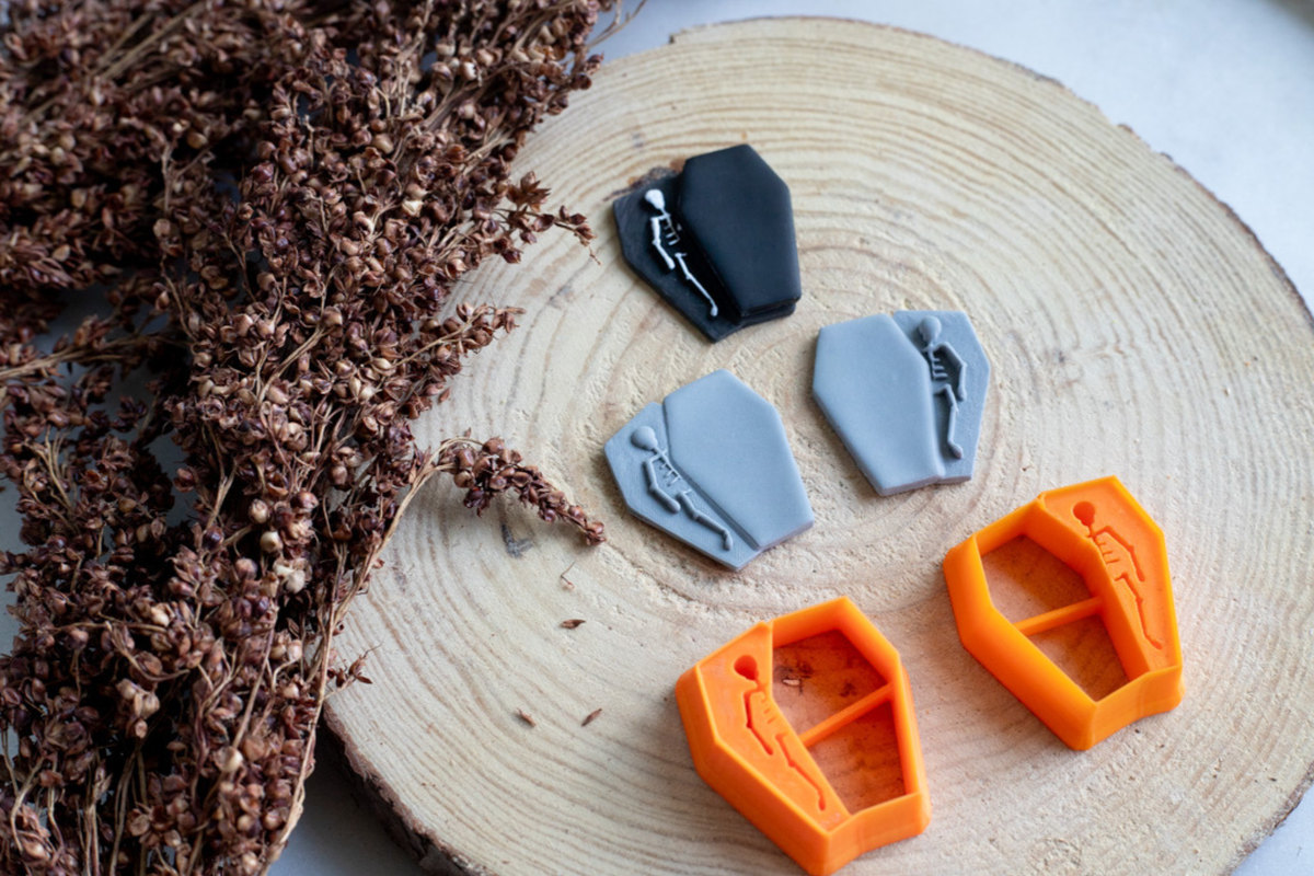 Four coffin-shaped cookie cutters in gray, black, and orange, each with a small skeleton detail, arranged on a round wood slice next to dried brown flowers.