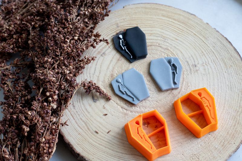 Four coffin-shaped cookie cutters in gray, black, and orange, each with a small skeleton detail, arranged on a round wood slice next to dried brown flowers.