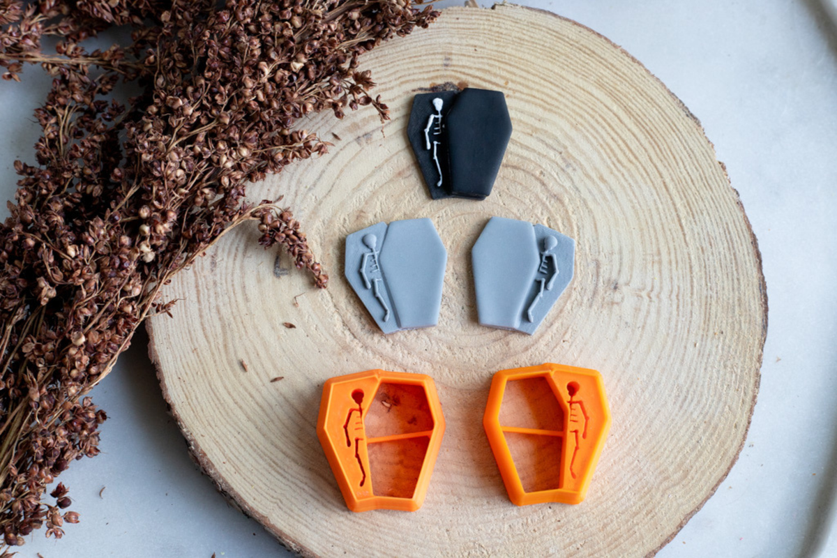 Four small coffin-shaped craft pieces and two matching orange molds arranged on a round wood slice with dried brown flowers beside it.